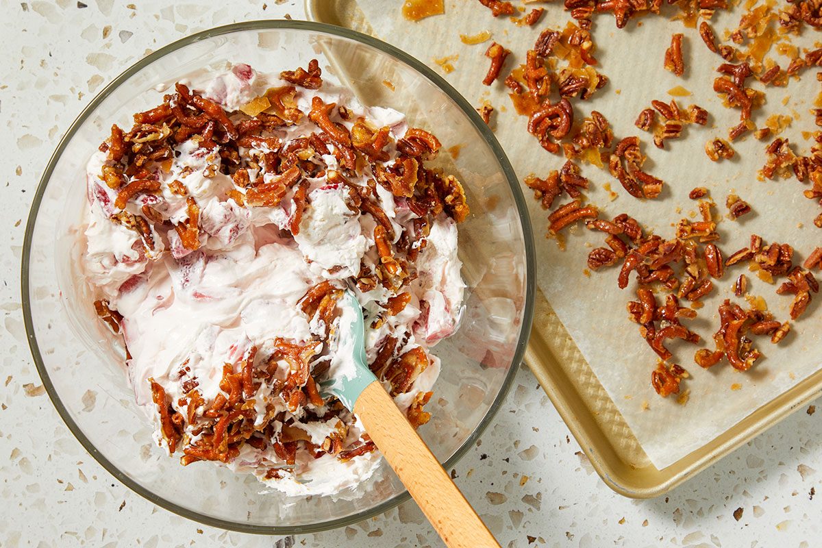 Overhead horizontal image of a glass bowl containing a creamy, pinkish fruit mixture topped with candied pecans, with a blue and wood spatula partially submerged. To the right, a gold baking sheet lined with parchment paper holds scattered glazed pecans.