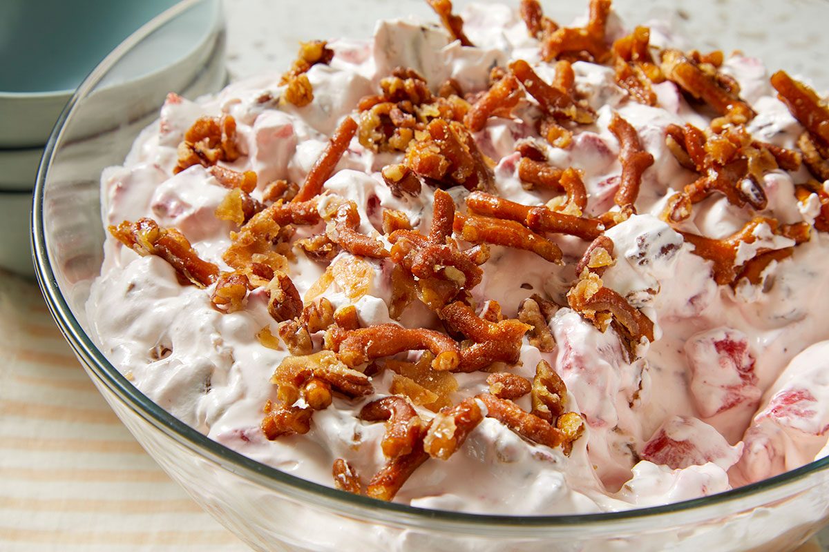 Eye Level horizontal image of a creamy pink and white dessert or salad in a clear glass bowl, featuring visible chunks of strawberries and marshmallows. It is generously topped with glazed pretzel pieces and chopped pecans.