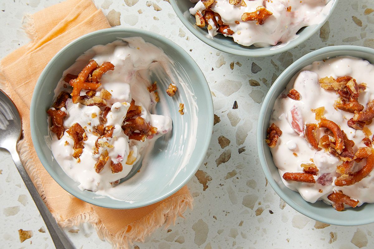 Overhead horizontal image of three light-colored bowls filled with a creamy, pale pink fruit salad or dessert. The dishes are generously topped with crunchy pretzel pieces and chopped nuts. A silver spoon and an orange napkin are visible on the left side of the frame against a mottled countertop background.