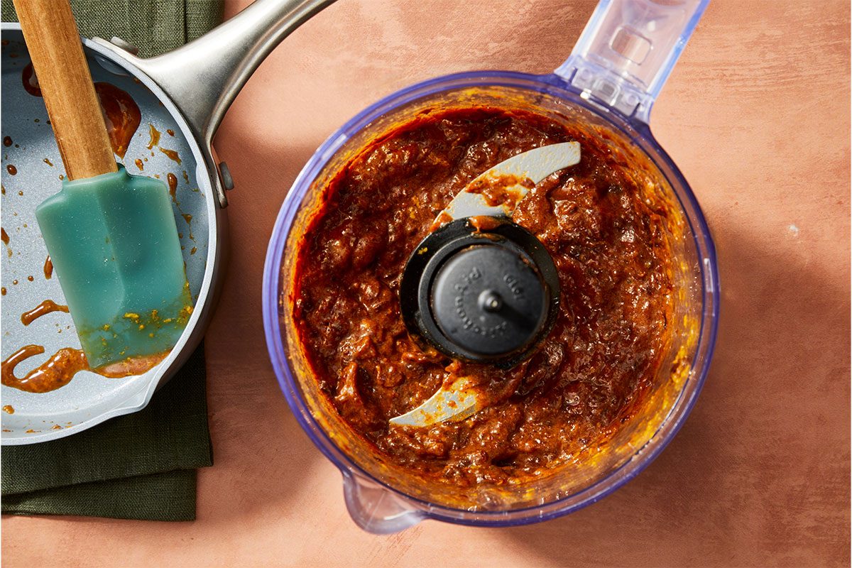 Overhead horizontal image of a food processor containing a sauce-like mixture with a spatula and pan visible to the side. The sauce is a dark red-brown color and sits within the food processor bowl, surrounding the metal blade. To the side, a pan contains residue of the sauce, and a spatula sits inside it. | [Original] RMS 281597; Sugar Plum Thumbprint Cookies; How to; Step 1; In a small saucepan; combine all jam ingredients. Cook; over medium heat; until prunes are softened; about 3 minutes. Remove from heat; cool slightly. Place jam in a food processor or high-powered blender; process until smooth; set aside.