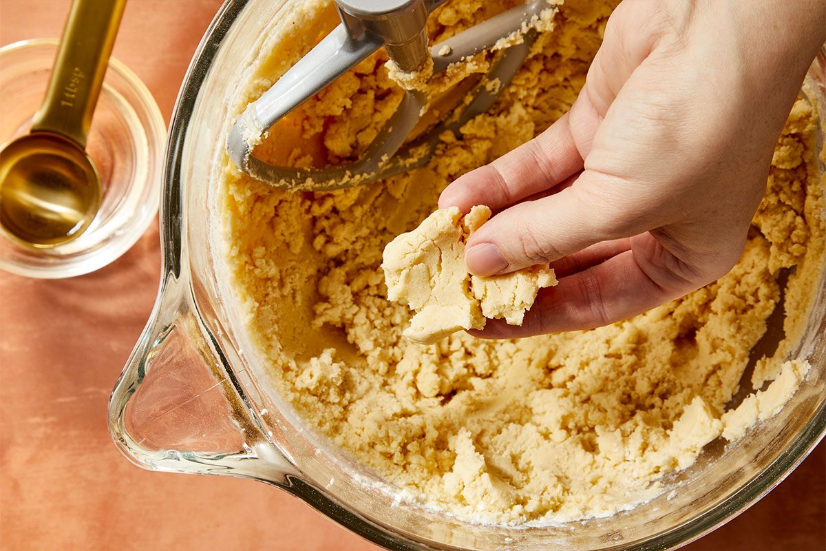 Eye Level horizontal image of cookie dough batter being held in someone's hand, after being scooped from a large clear glass bowl with a mixer attachment. A gold tablespoon measuring spoon is visible in the background. | [Original] RMS 281597; Sugar Plum Thumbprint Cookies; How to; Step 2; In a large bowl, cream butter and sugar until light and fluffy. Beat in egg yolk and vanilla. In another bowl, whisk flour and salt; gradually beat into creamed mixture. If mixture is dry, add water until dough holds together when pressed.