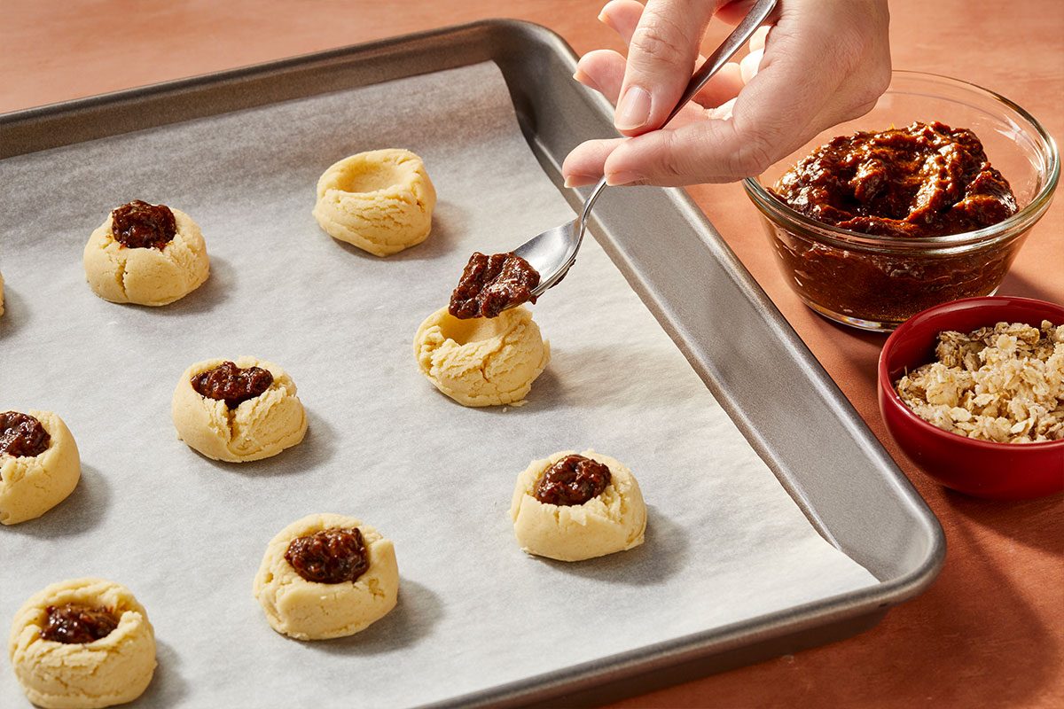 Eye Level horizontal image of cookies being filled with dark filling on a parchment paper-lined baking sheet. A hand holds a spoonful of the dark filling, with a small bowl of the filling and another bowl of oats visible in the background. | [Original] RMS 281597; Sugar Plum Thumbprint Cookies; How to; Step 3; Shape dough into 1-in. balls. Place 2 in. apart on parchment-lined baking sheets. Press a deep indentation in center of each with the end of a wooden spoon handle. Fill with prepared jam. Sprinkle crumble over jam on cookies