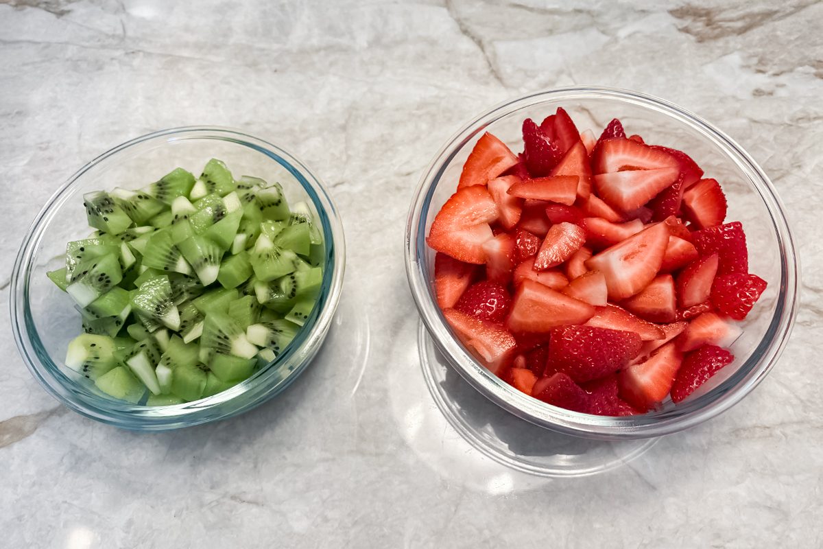 Two glass bowls on a marble surface; one bowl contains chopped kiwi, and the other contains sliced strawberries.