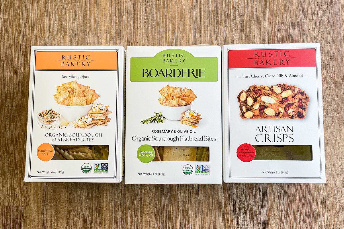 Three boxes of Rustic Bakery crackers are displayed side by side on a wooden surface. The flavors shown are Sourdough Flatbread Bites, Rosemary & Olive Oil Flatbread Bites, and Tart Cherry, Cocoa Nib & Almond Crisps.