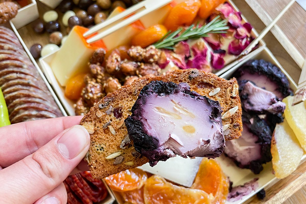 A hand holds a seeded cracker topped with a slice of purple cheese. In the background, there is a charcuterie board with nuts, dried fruit, crackers, and garnishes.