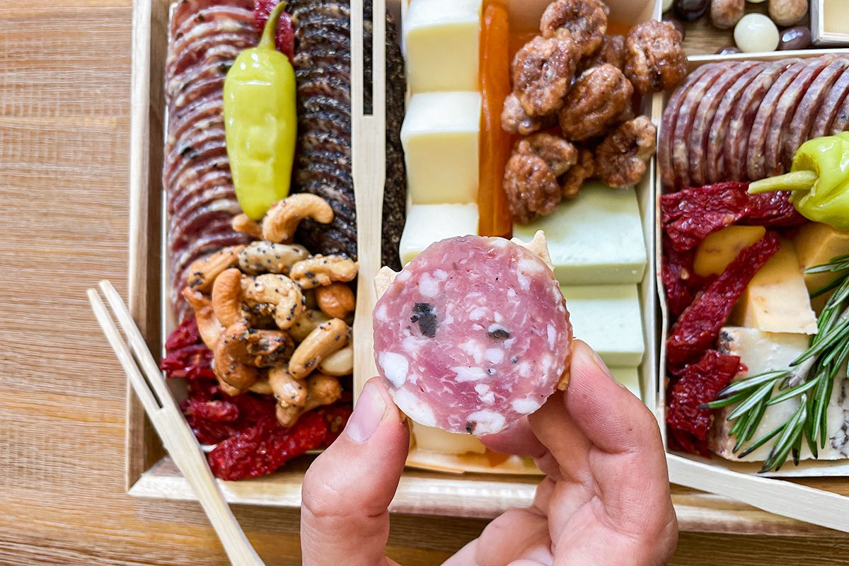 A hand holds a slice of salami over a charcuterie board filled with assorted meats, cheeses, nuts, pickled peppers, and garnishes on a wooden table.