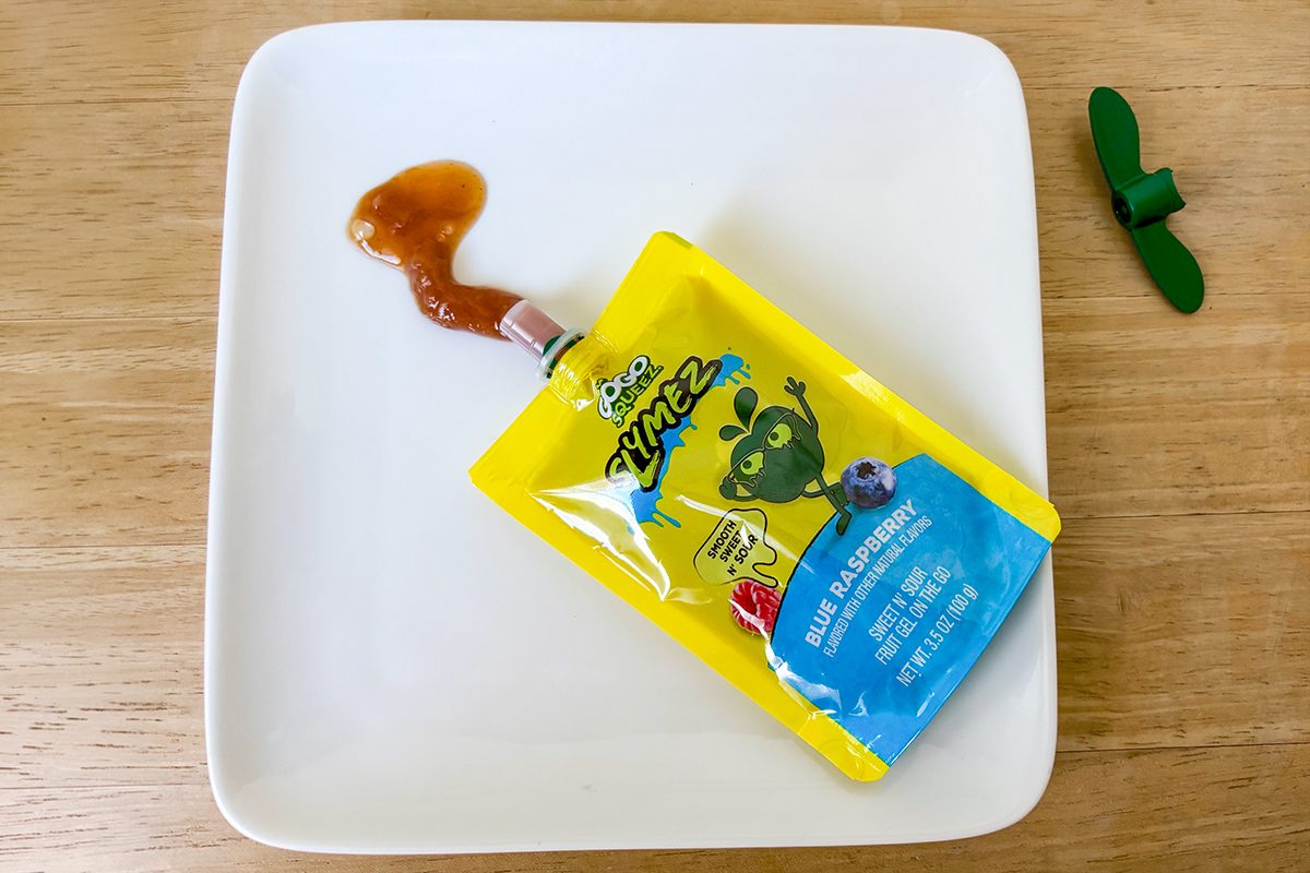 A yellow and blue snack pouch with a cartoon frog design lies on a white plate, squeezed open with brown sauce oozing out. A green plastic cap is placed nearby on the wooden surface.