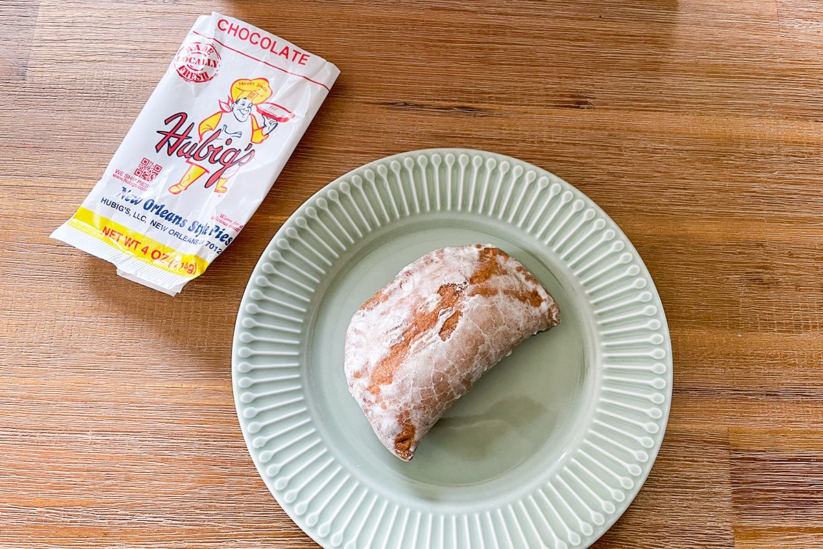 A chocolate halvah snack partially unwrapped from its packaging sits on a light green plate atop a wooden table. The wrapper is white with red and yellow accents and shows an illustrated character.