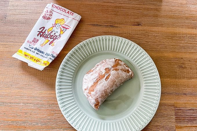 A chocolate halvah snack partially unwrapped from its packaging sits on a light green plate atop a wooden table. The wrapper is white with red and yellow accents and shows an illustrated character.