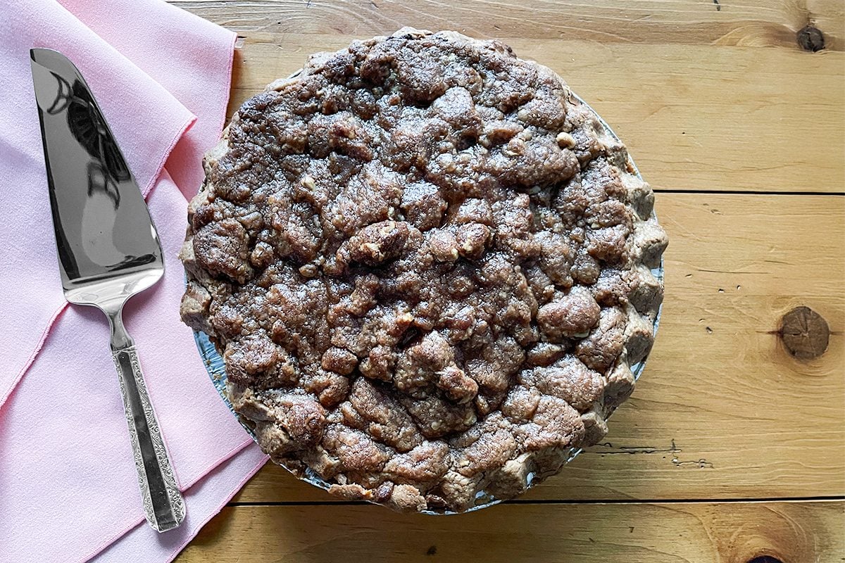 A crumb-topped pie sits on a wooden table next to a pink napkin and a metal pie server.