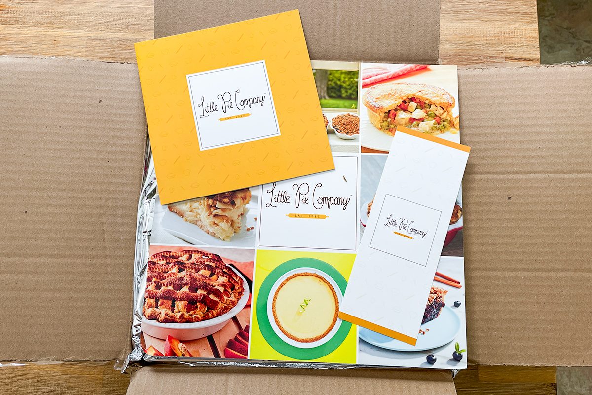 An open cardboard box contains a colorful food-themed package, a yellow square card, and a rectangular card, both labeled "Lake Pie Company." The package displays images of pies and other dishes.