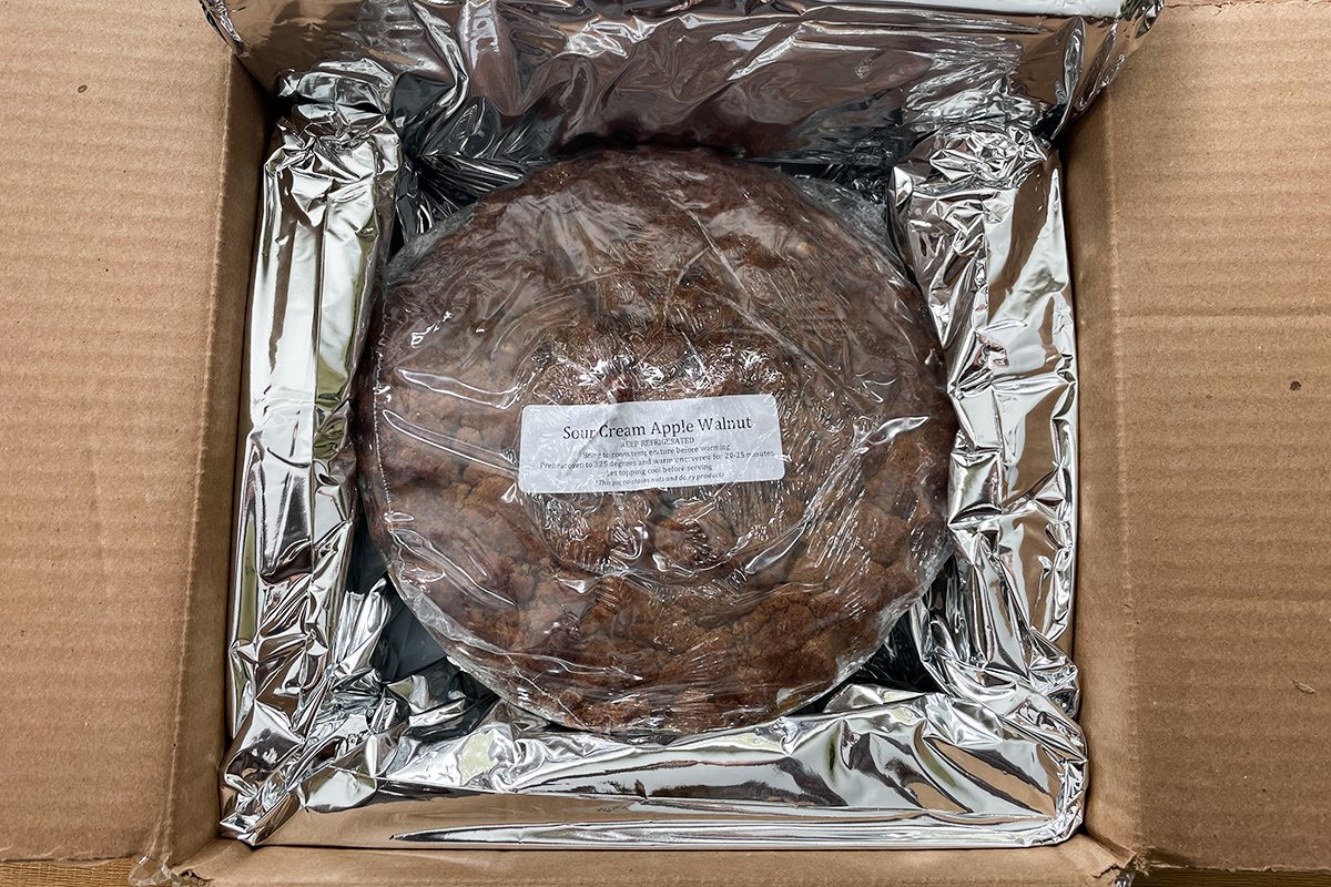 A round food item wrapped in plastic and placed inside a cardboard box lined with reflective foil. There is a white label on top of the wrapped item.