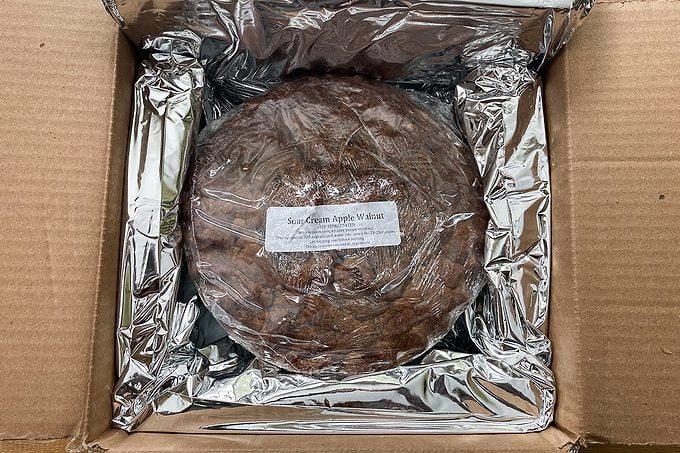 A round food item wrapped in plastic and placed inside a cardboard box lined with reflective foil. There is a white label on top of the wrapped item.