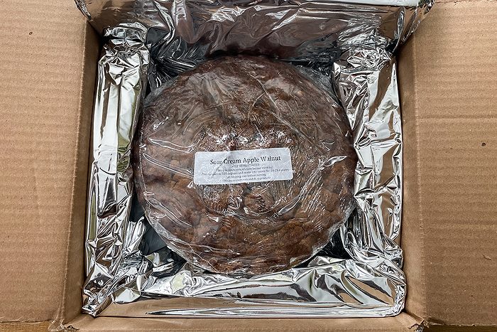 A round food item wrapped in plastic and placed inside a cardboard box lined with reflective foil. There is a white label on top of the wrapped item.