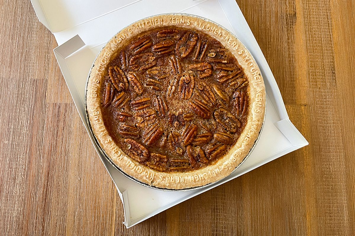 A pecan pie with a golden crust sits in an open white box on a wooden table.