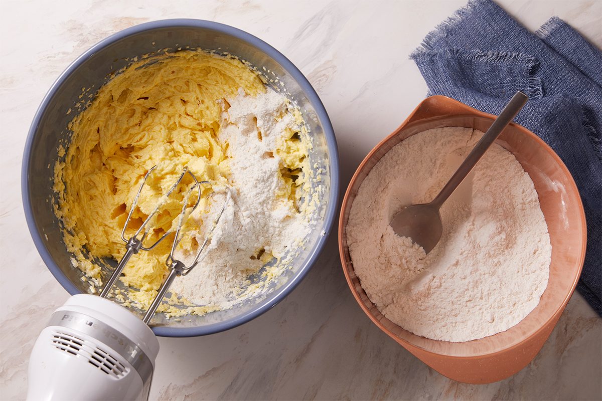 Overhead horizontal image of tea cake batter being mixed with an electric mixer in a blue bowl. Flour is being added to the batter, and a separate bowl containing more flour with a spoon is on the side.
