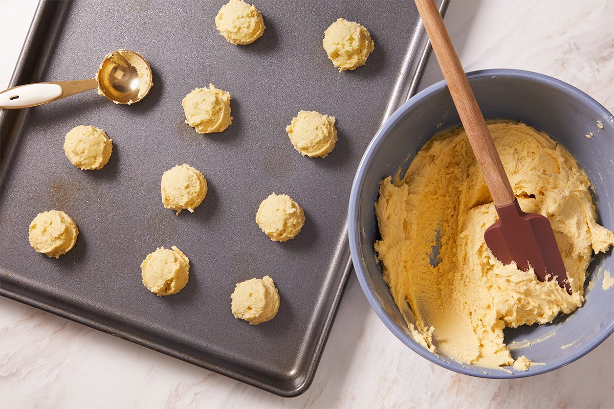 Overhead horizontal image of tea cake dough balls arranged on a baking sheet. A gold measuring spoon sits on the sheet next to some of the dough balls, with a light blue bowl of remaining batter and a brown spatula visible. The recipe calls for butter, sugar, eggs, vanilla extract, flour, baking powder, and salt.