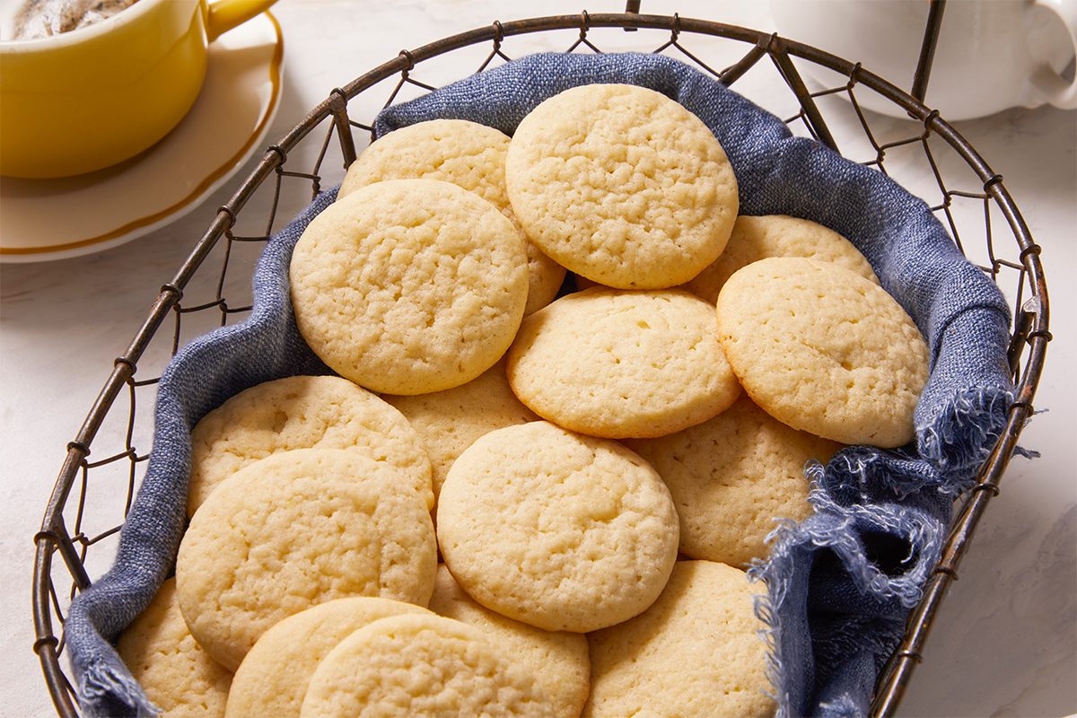 Eye Level horizontal image of tea cakes. A wire basket containing several tea cakes layered on a blue cloth. A mug is slightly visible in the background.