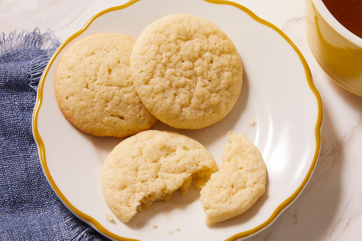 Overhead horizontal image of Tea Cakes on a plate with a bite taken out of one. The recipe is almost complete. Visible ingredients include flour, sugar, eggs, butter and vanilla extract.