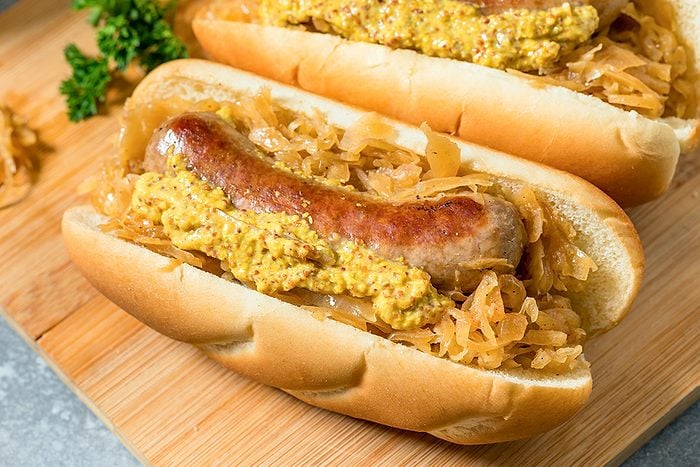 Two bratwursts in buns topped with sauerkraut and coarse mustard are placed on a wooden cutting board, with a sprig of parsley in the background.