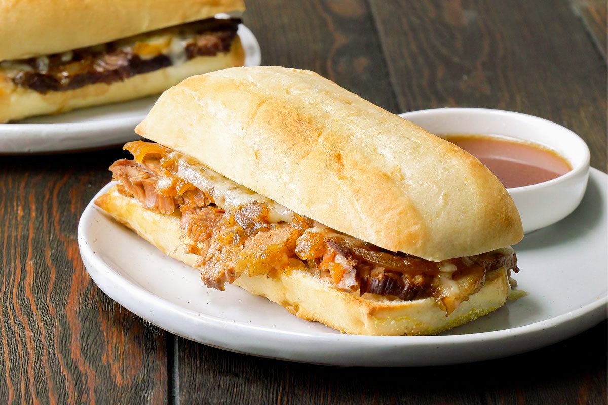A toasted sandwich filled with shredded meat and melted cheese sits on a white plate next to a small cup of dipping sauce. Another sandwich is partially visible in the background.