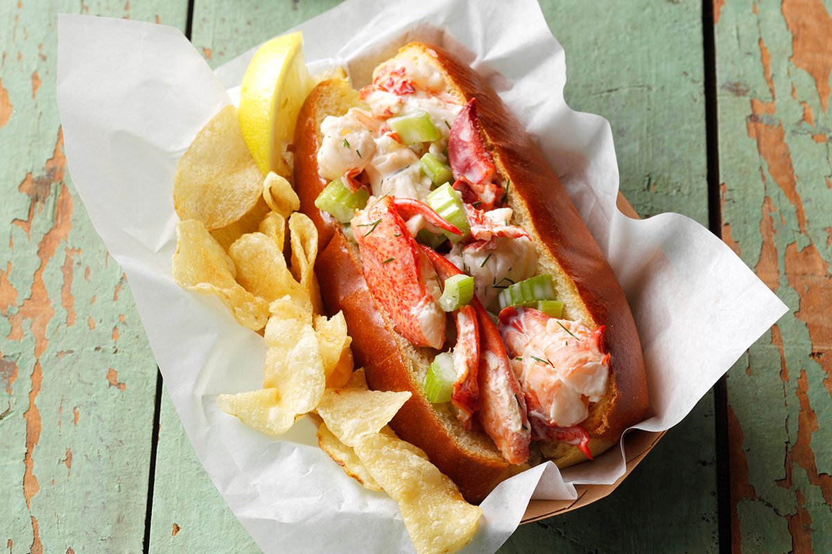 A lobster roll topped with diced celery sits in a paper-lined basket next to a pile of potato chips and a lemon wedge on a rustic green wooden table.