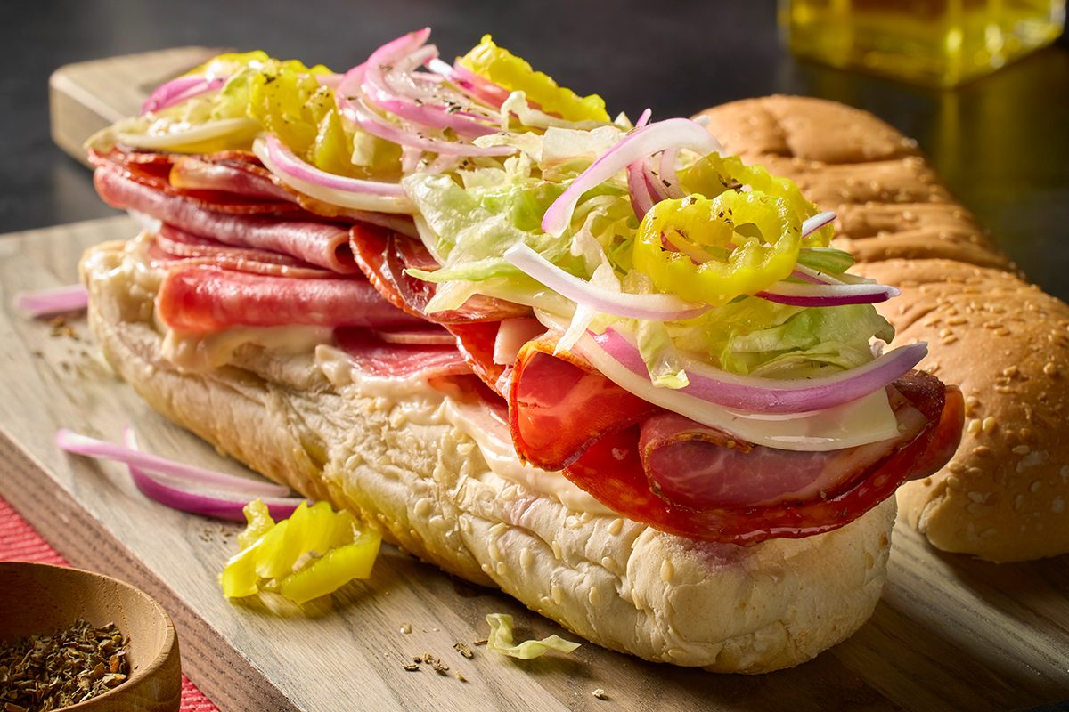 Eye Level vertical image of a fully assembled Italian sub sandwich overflowing with deli meats, cheese, shredded lettuce, sliced red onion, and pepperoncini. It rests on a wooden cutting board, with bottles of oil and vinegar, and a small bowl of oregano in the background.
