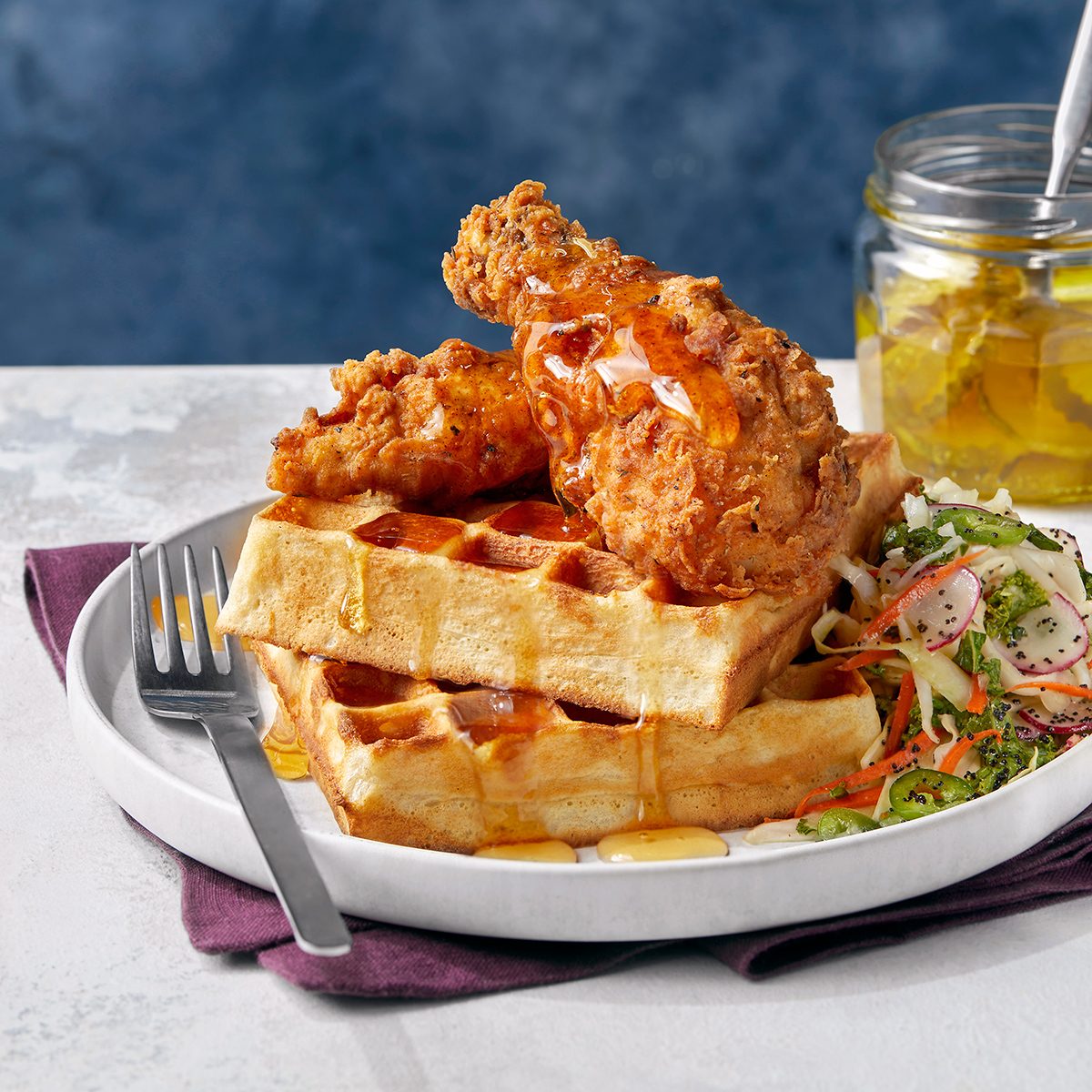 A plate with two stacked waffles topped with two pieces of fried chicken, drizzled with syrup, served with a side of coleslaw. A fork rests on the plate; a jar of pickles is in the background.