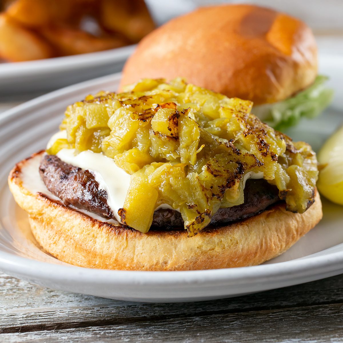 A hamburger on a white plate, topped with melted cheese and chopped green chilies, with a bun top slightly off to the side. A pickle spear and another plate of food are visible in the background.
