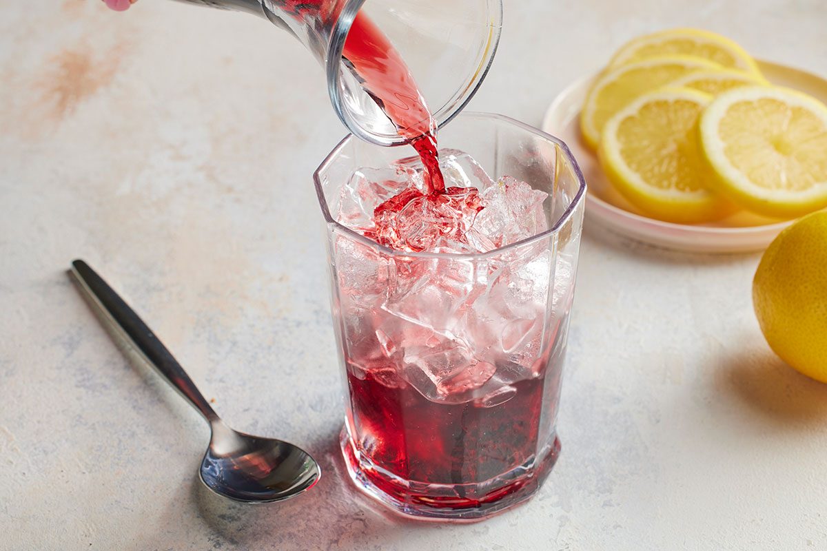 Eye Level horizontal image of red liquid being poured from a glass pitcher into a clear glass filled with ice. A plate of lemon slices, a whole lemon, and a spoon are visible in the background.