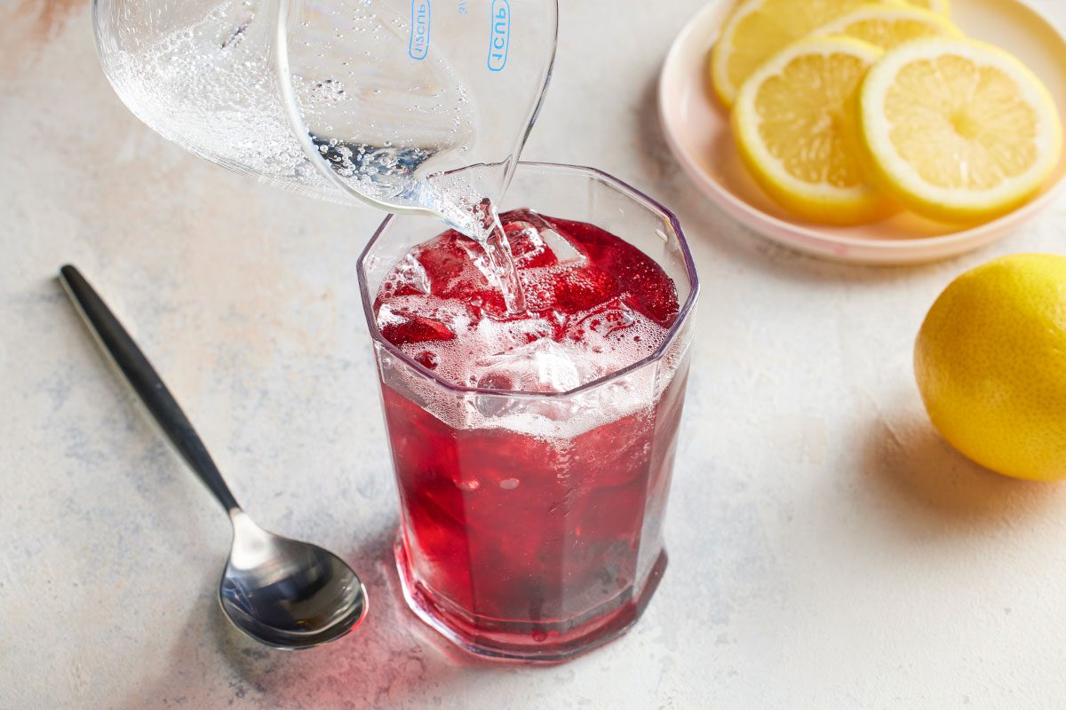 Eye Level horizontal image of sparkling water being poured from a glass measuring cup into a glass filled with a dark red beverage and ice. Sliced lemons sit on a plate in the background, along with a whole lemon and a spoon.