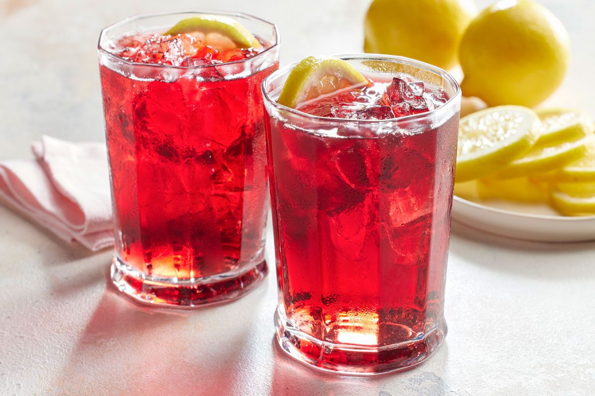 Eye Level horizontal image of two filled glasses with a red liquid and ice, garnished with a lemon slice. Lemon slices are also piled onto a plate in the background.