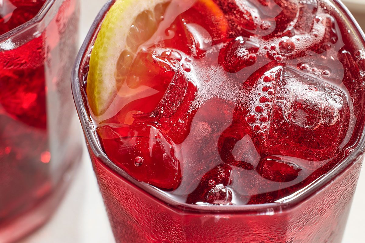 Eye level horizontal image of a beverage in a glass. The glass contains ice and a slice of lemon, with visible condensation on the outside.