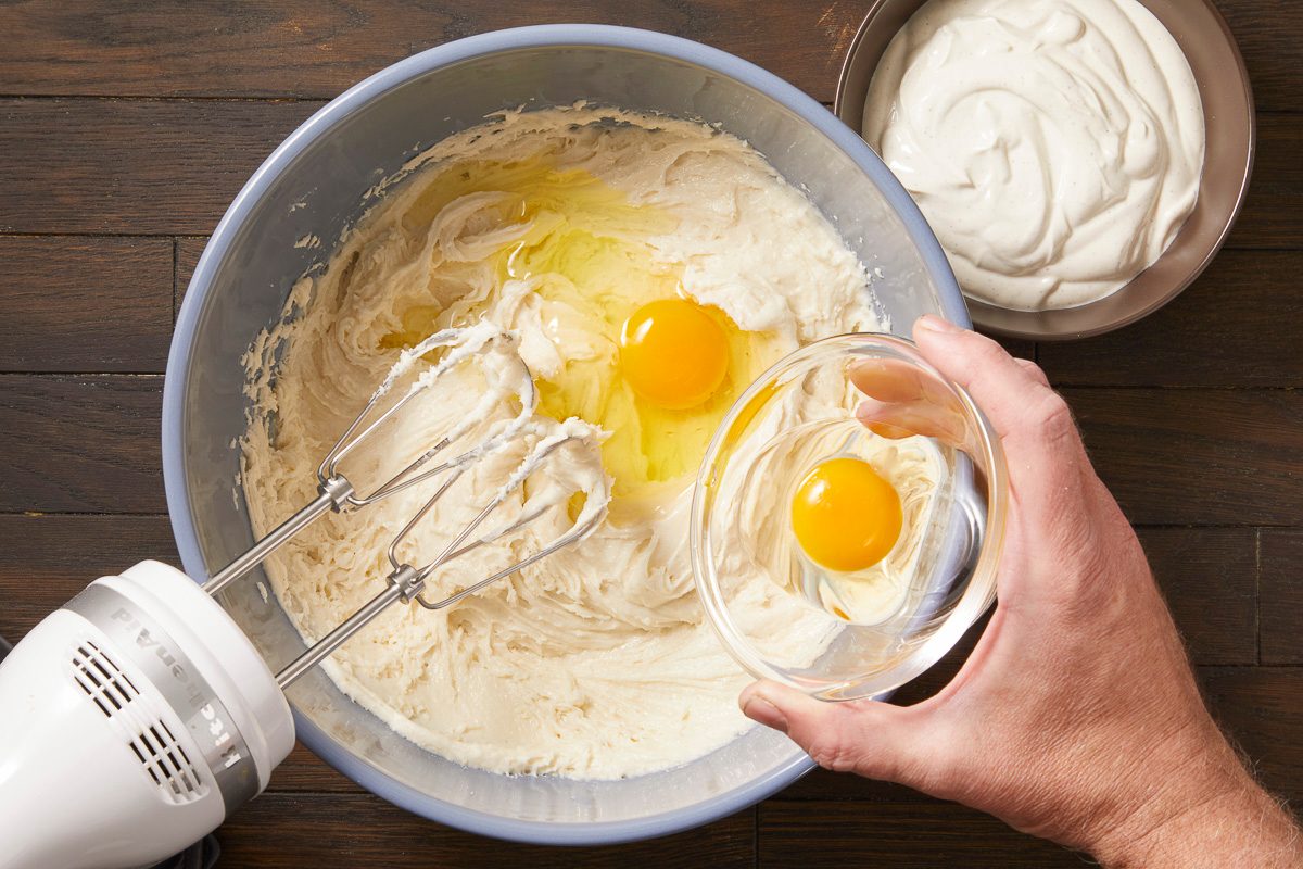 Overhead horizontal image of a hand adding an egg from a small glass bowl into a large blue mixing bowl that already contains creamy batter and two other eggs. An electric KitchenAid hand mixer rests in the batter, with a bowl of sour cream visible in the background.