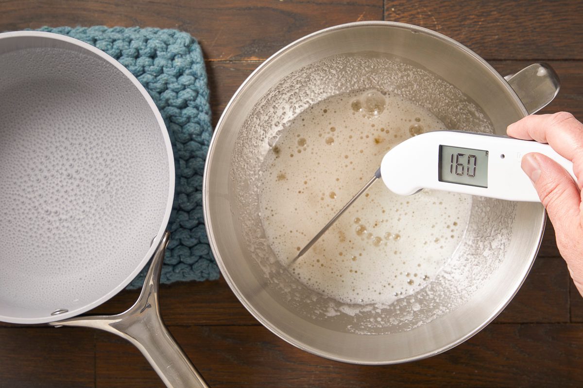 Overhead horizontal image of a hand holding a digital thermometer in a stainless steel pot containing a frothy liquid. The thermometer displays a reading of '160', indicating a temperature measurement. A second pot with a white interior rests on a blue mat to the left, all on a wooden surface.