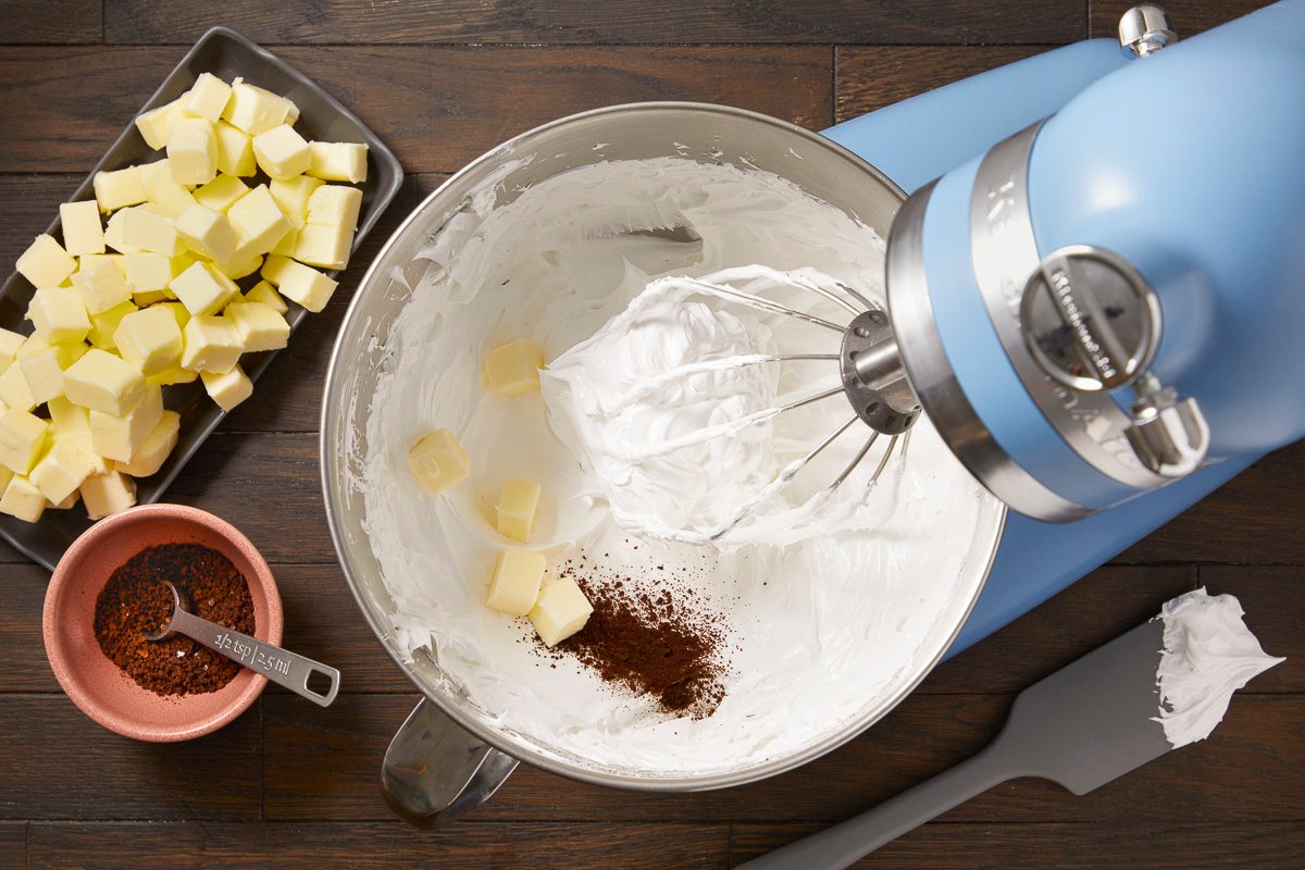 Overhead horizontal image of a light blue stand mixer bowl containing white frosting, butter cubes, and a small mound of instant coffee powder. A whisk attachment is immersed in the frosting, and additional cubed butter is on a rectangular plate nearby. A small pink bowl holds more instant coffee powder with a measuring spoon.