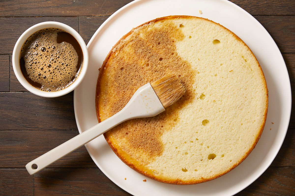 Overhead horizontal image of a pastry brush applying a liquid to a round cake layer. A portion of the cake is already moistened, while another part remains dry. A small white bowl filled with coffee is visible next to the cake.