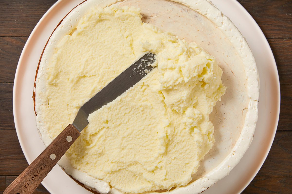 Overhead horizontal image of a cake being frosted. A generous amount of light yellow frosting is being spread over the top of a cake layer using an offset spatula, with a white frosting border already present around the cake's edge.