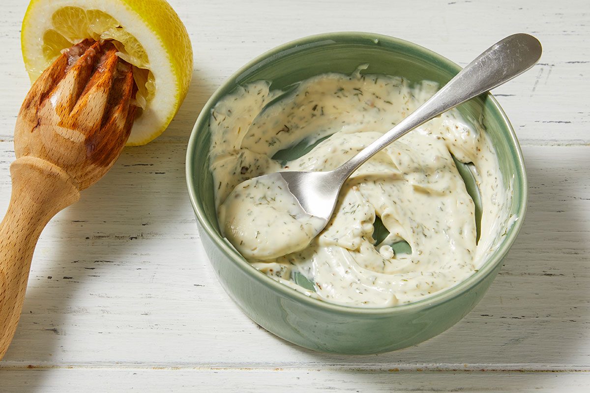 Overhead horizontal image of a green bowl filled with dill mayonnaise and a silver spoon. A yellow lemon is being squeezed by a wooden reamer on a white distressed wooden surface, adding fresh lemon juice to the mixture.