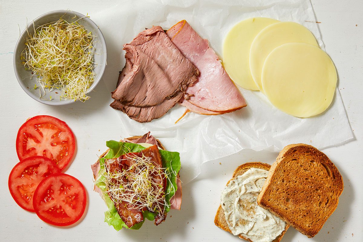 Overhead horizontal image of a sandwich being assembled, with various ingredients laid out on a white surface. One half of a toasted club sandwich is partially built with lettuce, bacon, and alfalfa sprouts, while a separate slice of toasted whole wheat bread is spread with dill mayo. Beside them are bowls and parchment paper holding alfalfa sprouts, deli roast beef, deli ham, provolone cheese slices, and sliced tomatoes.