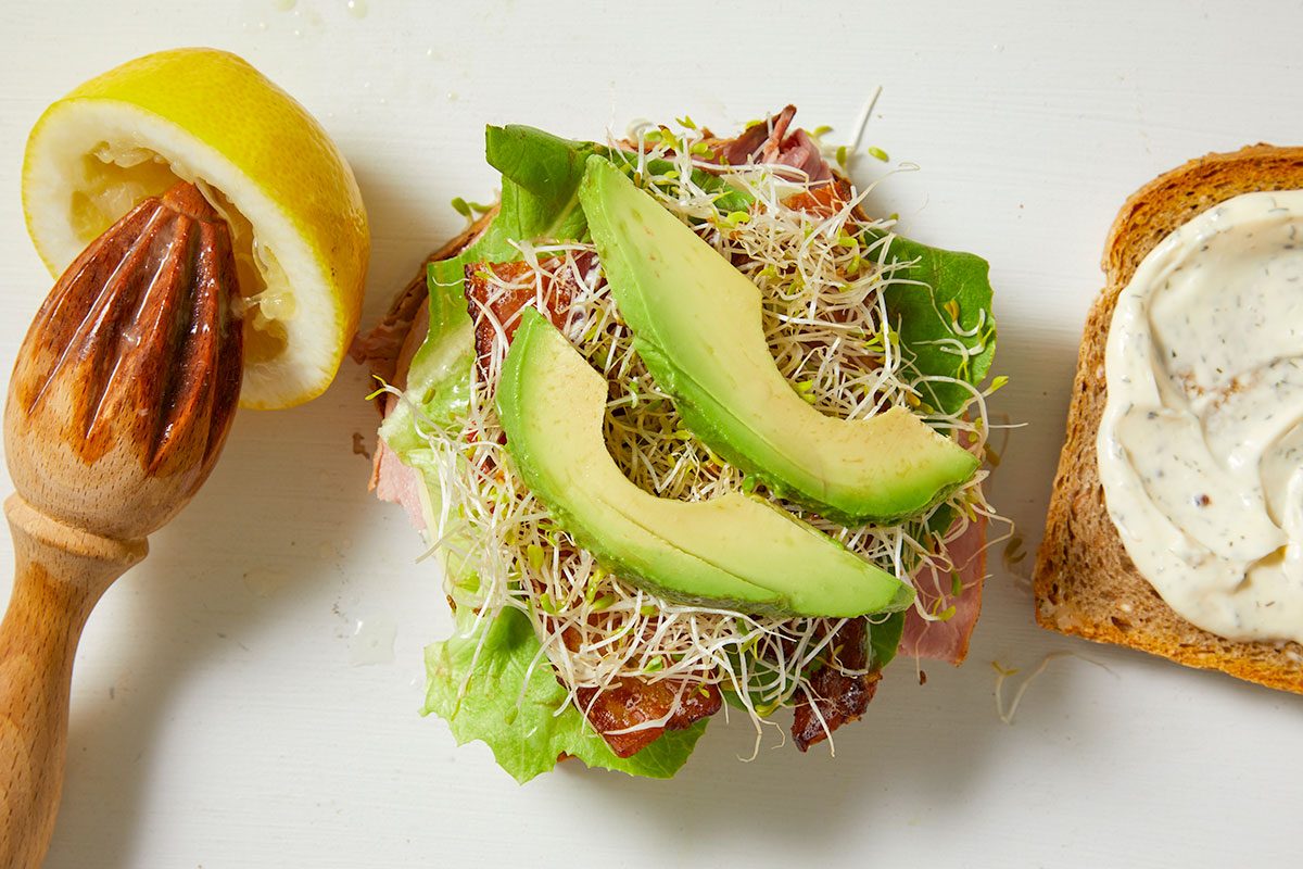 Overhead horizontal image of a lemon being squeezed with a wooden reamer, with droplets of fresh juice visible on the white surface. To the right, a slice of toasted whole wheat bread is spread with dill mayo. In the center, an open-faced sandwich base is topped with Bibb lettuce, deli meats, alfalfa sprouts, avocado slices, and crumbled bacon.