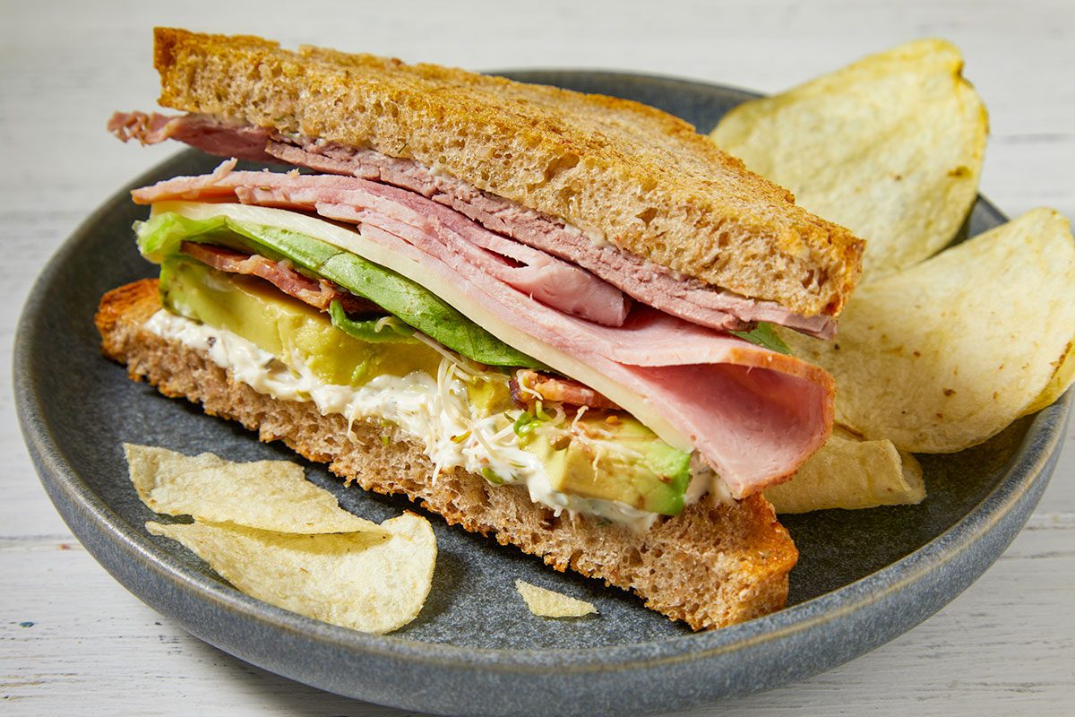 Eye Level horizontal image of a toasted club sandwich, cut in half and showcasing its bountiful layers, resting on a gray plate with potato chips. The sandwich features toasted whole wheat bread filled with roast beef, ham, provolone cheese, Bibb lettuce, sliced avocado, crumbled bacon, alfalfa sprouts, and a creamy dill mayonnaise.