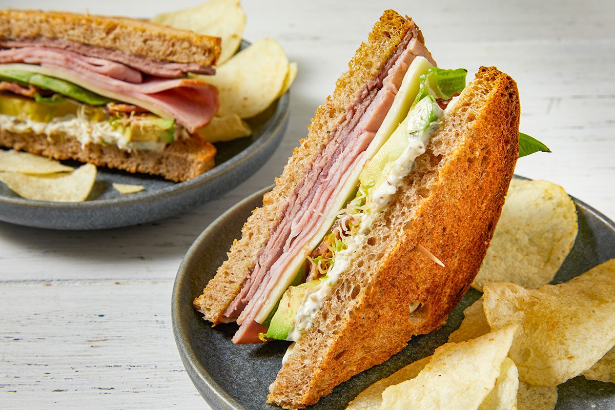 Eye Level horizontal image of two hearty Toasted Clubs with Dill Mayo sandwiches. The foreground sandwich is cut in half and stands upright, showcasing layers of deli roast beef, ham, provolone, Bibb lettuce, avocado, alfalfa sprouts, and a creamy dill mayonnaise on toasted whole wheat bread. Potato chips are scattered on the plates as a side.