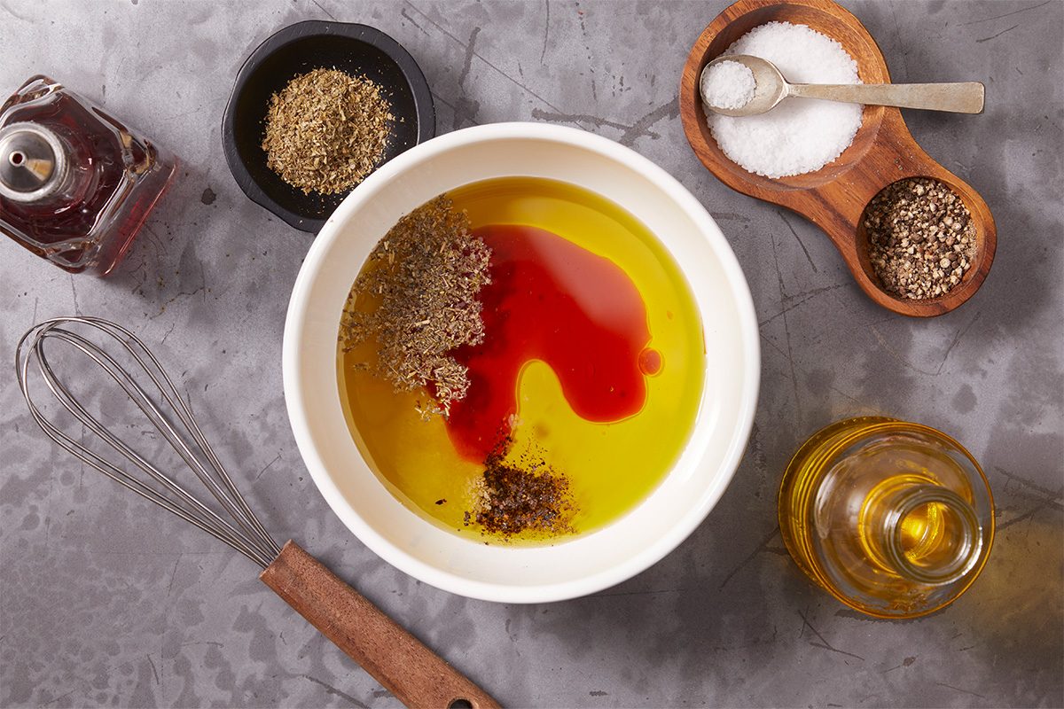 Overhead horizontal image of a white bowl containing olive oil, red wine vinegar, dried herbs, and black pepper, with salt in a wooden dish to the side. A whisk, a bottle of red wine vinegar, and a bottle of olive oil are also visible on the gray counter.