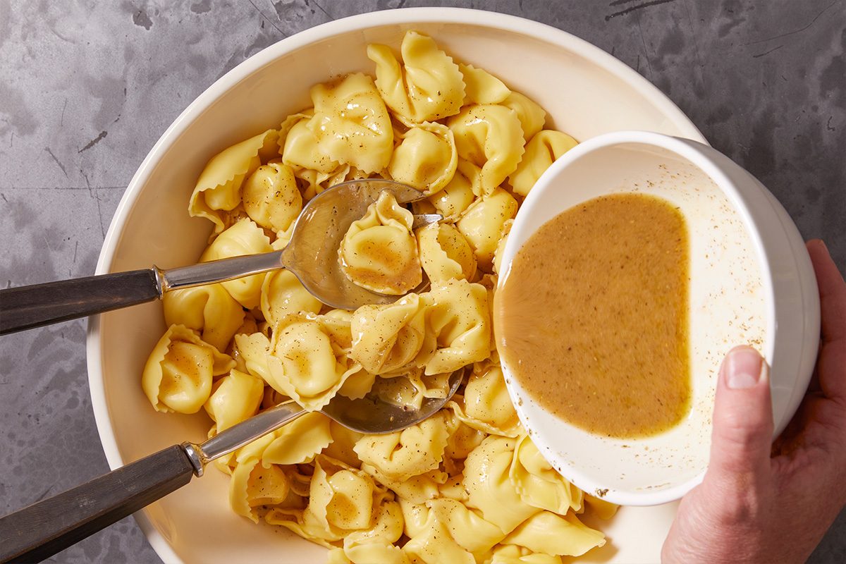 Overhead horizontal image of a hand pouring a light brown dressing from a white bowl over cooked tortellini in a cream-colored serving bowl. The tortellini is lightly seasoned with visible specks of pepper or herbs, and two serving spoons rest within the pasta, suggesting a mixing step.