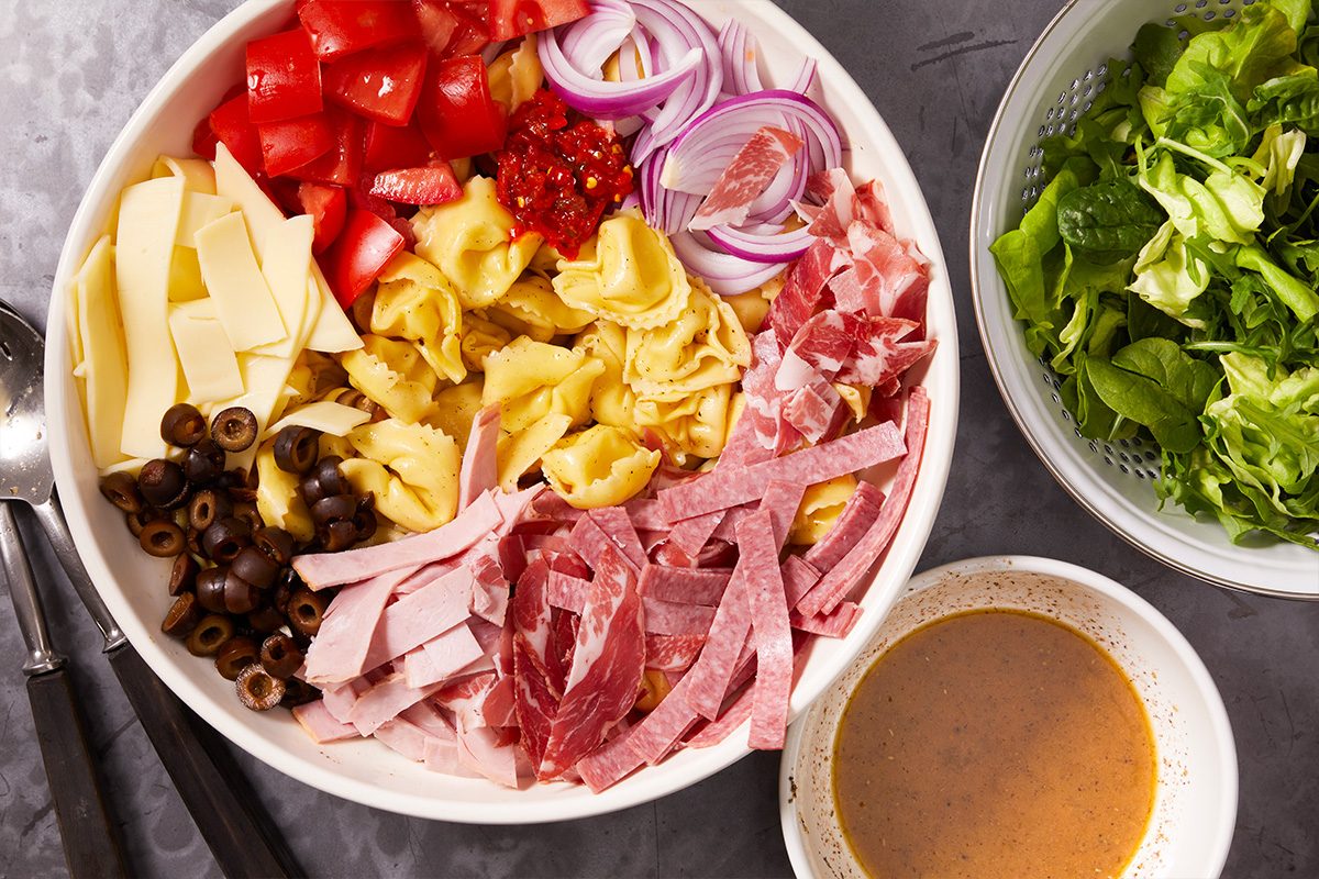 Overhead horizontal image of a large white bowl filled with various salad ingredients, including tortellini, diced tomatoes, sliced red onions, a dollop of red relish, sliced cheese, sliced black olives, sliced ham, prosciutto, and salami strips. A colander with mixed green lettuce and a small bowl of dressing are placed alongside the main bowl on a dark gray surface.