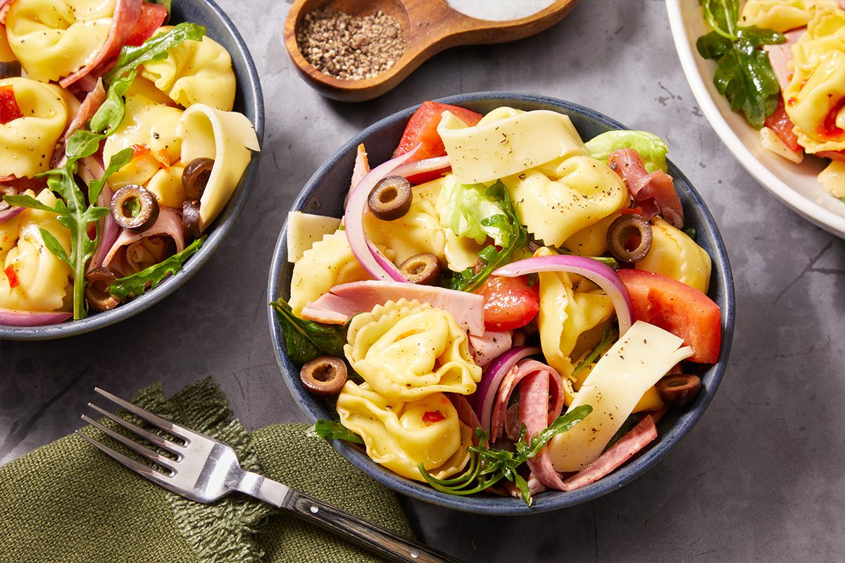 Overhead horizontal image of two blue bowls filled with a vibrant pasta salad on a gray surface. The salad features tortellini, slices of deli meat, red onion, black olives, chopped tomatoes, green arugula, and sliced cheese. A fork rests on a green napkin, with salt and pepper in wooden spoons in the background.