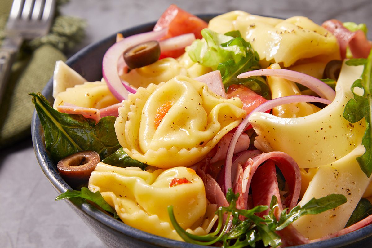 Eye Level horizontal image of a vibrant pasta salad served in a dark blue bowl. The salad features cheese tortellini, sliced red onion, black olives, chopped tomatoes, fresh spinach, arugula, and slices of ham and salami, all coated in a light dressing.