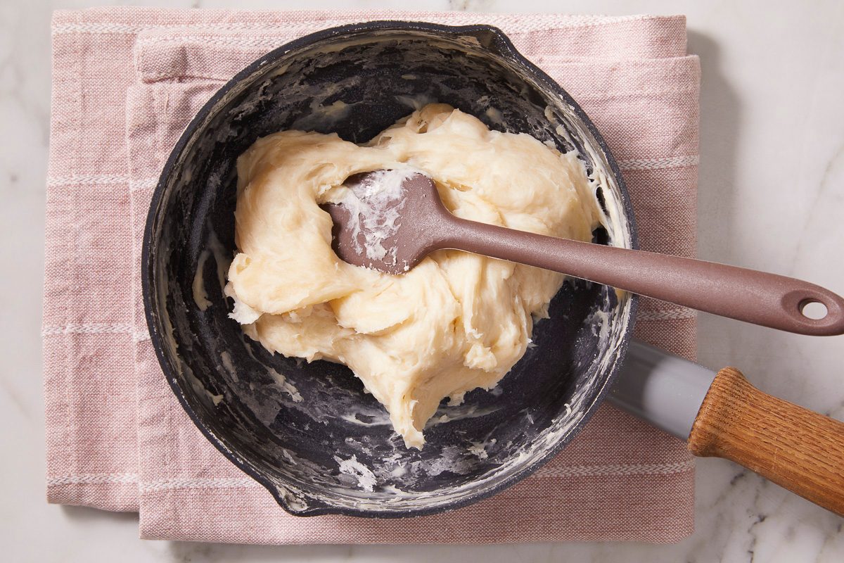 Overhead horizontal image of a thick, light-colored flour mixture being stirred with a brown spatula in a dark saucepan. The saucepan rests on a light pink striped kitchen towel, set on a white marble surface.