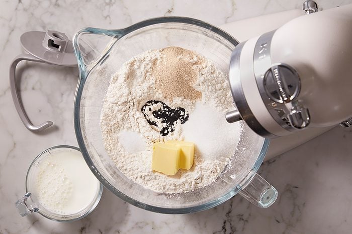 Overhead horizontal image of a stand mixer bowl filled with dry ingredients for ube bread. White flour, sugar, yeast, salt, softened butter pats, and a dark blob of ube extract are visible inside the bowl. A glass measuring cup containing whole milk sits beside it.